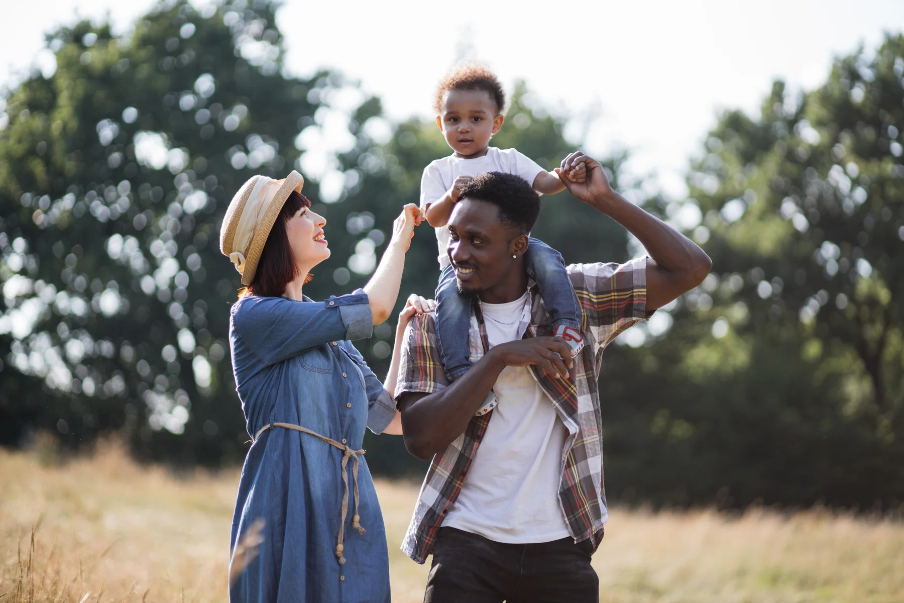 A father with his young son on his shoulders, smiling with his wife in a sunlit meadow
